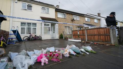 A cordon outside the front of the property with a police car parked in the road. Flower tributes have been left on the driveway of the house