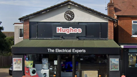 Exterior of a Hughes shopfront. Washing machines and other white appliances can be seen in the shop window. Written on a green awning is the phrase, the electrical experts.
