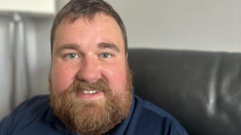 Kevin Clemitson is sitting on a black leather couch. He has mid brown hair and a lustrous beard and moustache and is fair skinned with freckles. He is smiling and wearing a navy blue top. 