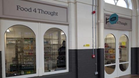 The front of the shop has two big cylinder shaped windows. The walls are brick and are painted half white and half black. You can see trolleys inside with food items and others on it. 