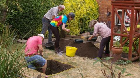 A view of four people digging a hole in a garden. One man in a pink t-shirt is inside a square hole, while three other people look through a pile of dirt.