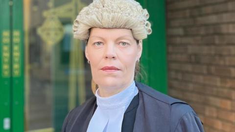 Claire Davies KC stands outside a court entrance wearing her black gown with white collar and her barristers wig.