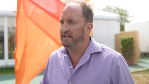 Guto Harri is facing sideways to the camera, wearing a lavender shirt, stood on the Hay Festival grounds.