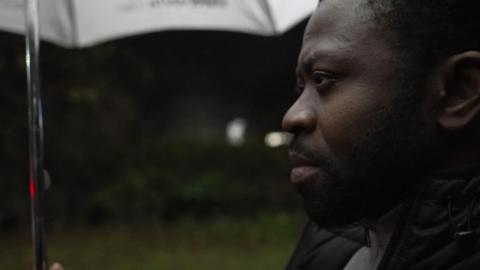 A close up of Mr Morakinyo, who is looking serious, wearing a black coat and holding an umbrella.