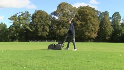 A man pushes a black lawnmower across a green sports field. Trees are in the background.