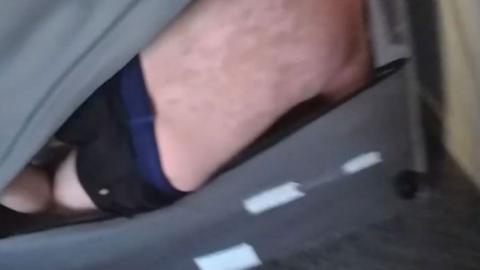 A blurry police-cam image of a man's pale back and dark pants. He is tucked into the back of a sofa with his knees raised. The grey fabric is parted to show his back