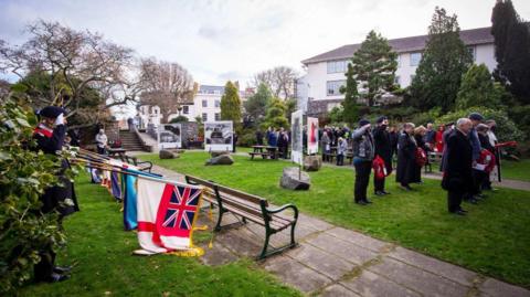 People stood in a park during a military memorial service. A group of people on the left-hand side are holding flags on poles which have been lowered to the ground. Other people stood on a patch of grass have poppy wreaths in their hands.