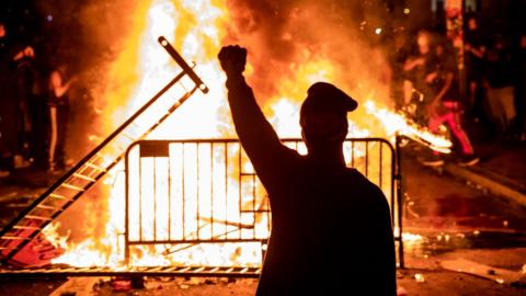 A protester stands in front of a flaming barricade holding a fist in the air.