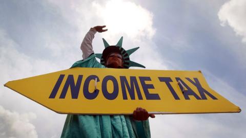 A man dressed at the Statue of Liberty holding a sign saying 'income tax'