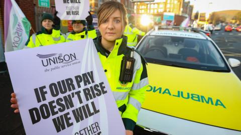 A paramedic on strike holds a placard that reads: IF OUR PAY DOESN'T RISE WE WILL