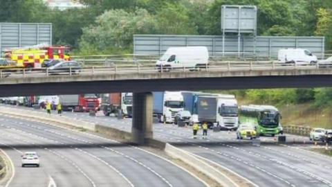The M1 motorway, showing a bridge, and several lanes of motorway below. There are emergency services at the scene, one car on the southbound section, and a fire engine on the bridge. Other vehicles are driving over the bridge. 
