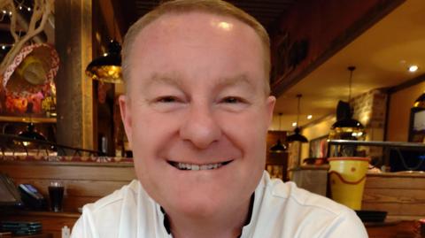Michael Booth has short brown hair and a white top. He is smiling. He is sat in a restaurant. 