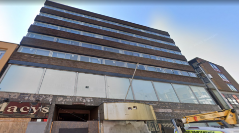 A large, box-shaped 1960s building, with rows of windows separated by brown brick sections. The ground floor is boarded up and looks derelict.