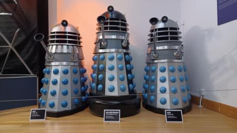 A group of three prop Daleks, conical robotic alien creatures, from the BBC science-fiction series Doctor Who, on display as museum exhibits against a white background. The two on either side are silver with blue trim, while the one in the centre stands slightly taller on a large black fender, with black stripes and a black dome section at the top in addition to the silver and blue of the others.