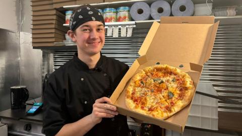 Joshua Steer holding up a pizza and smiling at the camera. He is wearing a black button up chefs jacket and a bandana