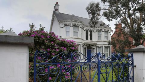 The former Kincora Boy's Home - a large, grey Victorian villa with bay windows on either side of its blue front door. The house is sited in a garden, behind a grey wall and decorative blue wrought-iron gates. There is a purple rhododendron plant in full blook in the garden. 