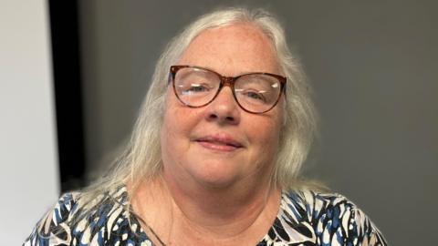 Jane Johnson, who has shoulder-length grey hair, wears brown glasses. She is photographed wearing a patterned white, dark blue and black top.