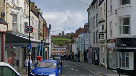 A street with shops on both sides and cars parked down one side. In the distance, at the end of the street, is a building and then a green hill with a ruined abbey at the top.