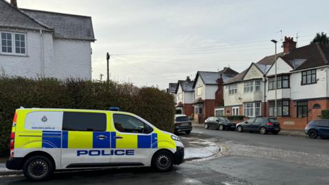 A police van parked on the side of a road surrounded by homes on a quiet residential street