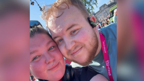A close up selfie of Richard with his girlfriend, Sophie at the Race for Life running event. Sophie, on the left, has brown hair and wears sunglasses on the top of her head. Richard is standing next to Sophie with his cheek touching her cheek. He wears a green t-shirt, a pink medal, earphones and has short blonde hair. 