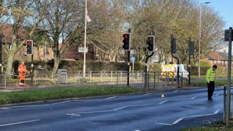 A police officer combs a cordoned off street for clues. The asphalt is wet and there is a single, yellow marker cone in the road.