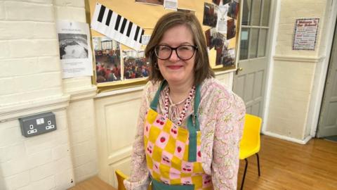 A woman wears a pink floral top, a colourful apron and glasses stands smiling in a school lunchroom. 