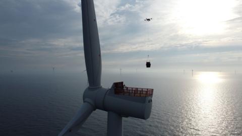 A drone carrying a box to a wind turbine in the sea. The sky is blue with sun shining through the clouds.
