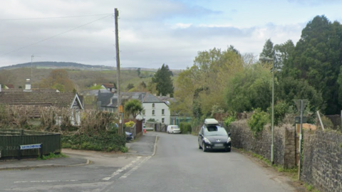A narrow road in a village. A car is travelling up the road. A van is parked further down the road. A large white property can be seen at the bottom of the road and is surrounded by trees and greenery. A stone wall lines one side of the street.