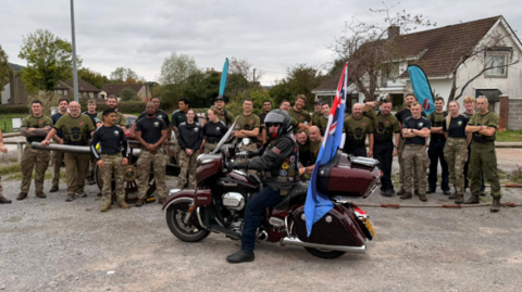 About a dozen military personnel, mainly men, in combat trousers surrounding the gun. There is also a man in the foreground on a vintage burgundy motorcycle