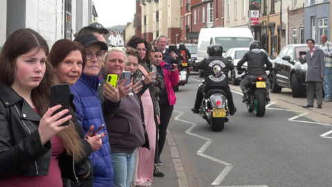 A number of people lining the street, many have phones which they are using to film the procession. There are motorbikes in the background, as if they have just driven past as part of proceedings. 