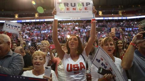 a woman in a USA T-Shirt holds a Trump sign as she stand at the front of the crowd, 10 August 2016