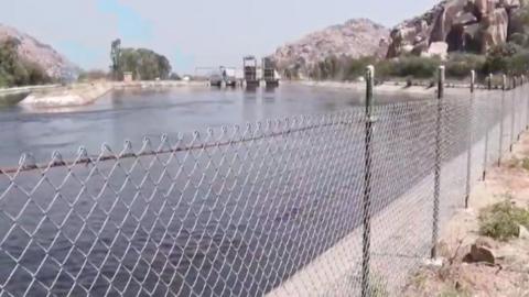 Blue water flows in the Sanapur river canal near Hampi. We can see a wire railing supported by metal pipes and boulders in the background. 