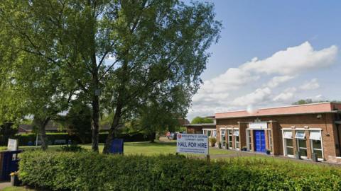 A community centre next to large trees on sunny day. Sign outside says "Hall for Hire". 