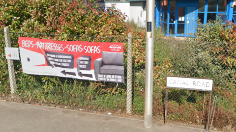 A red and white sign advertising beds, mattresses and sofas pinned to a wire fence outside. There is greenery behind it and a road sign next to it which says 'Canal Road'.