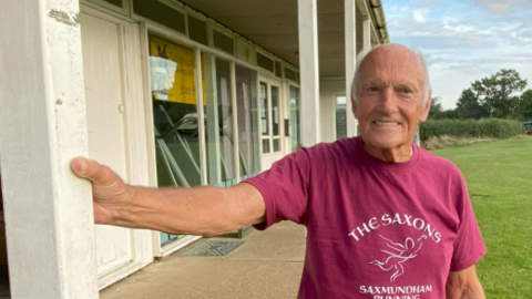 Gordon Merfield smiles at the camera as he rests one hand on a white beam on the exterior of a sports pavilion. He has some grey hair and wears a dark purple T-shirt with the Saxons running club logo on it. There is a grass sports pitch to the right of him.