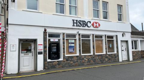 The front of the HSBC branch in St Sampson's. It has five widows on the front with the far left one having an ATM in it. There are two doors on the right and left hand side of the windows with the one on the right having a circular window above. The HSBC Logo sits above the windows in a central location.