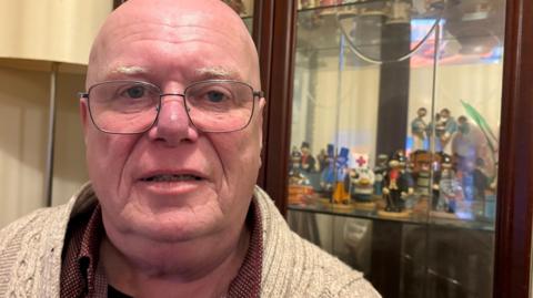 Andy Watson with very short hair and glasses looking at the camera while wearing a beige cardigan over a red shirt. There is a china cabinet behind him with various figures inside and a lamp with a light brown shade.