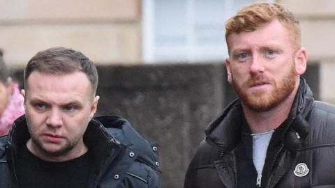 Tight head shot of two men walking outside a court building. One has brown hair, combed to the side, and the other has red hair and a beard.
