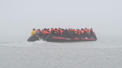 A small boat in the distance on the Channel. The boat is turned away from the camera, as migrants sit on either side of the boat, onwards to the UK.