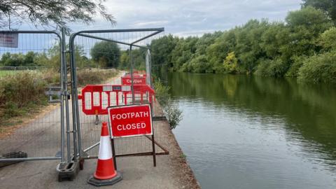 A footpath closed sign, wire fencing and a traffic cone mark the start of a path closure beside a river.
