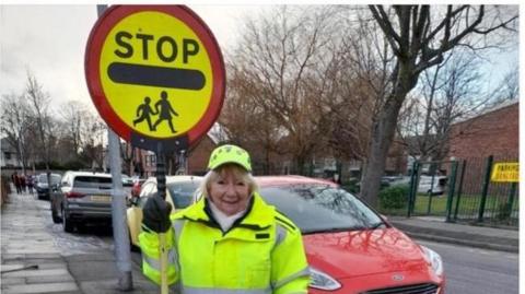 Margie is holding a stop sign and is dressed in full high visibility yellow school crossing patrol uniform. She stands on a pavement near a row of parked cars outside a school gates.