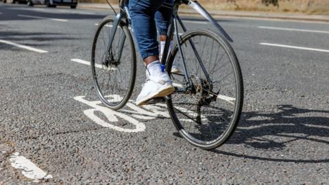 A road surface with a cycle lane marked out by a white bicycle symbol. A dark bike in the cycle land. All that can be seen of the cyclist is their legs and feet. They are wearing jeans and white trainers.
