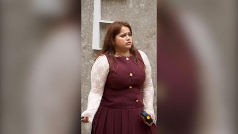 A woman in a burgundy dress with lace sleeves stands looking confused