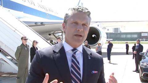 Pete Hegseth in a suit with a plane in the background.