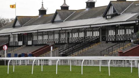 A general view of Musselburgh Racecourse. The grass in green and a white racetrack perimeter fence is surrounding it. In the background is the stand, which is white and black with grey standing areas.