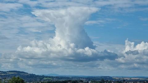 A huge cumulonimbus cloud builds high into the sky above the edges of a landscape of hills, fields and roof tops