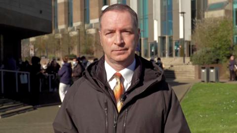 A man in a shirt and tie and rain jacket looking towards the camera