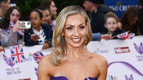 Amy Dowden stands outside in the sunshine wearing a mac and looking happy. She has long, blonde hair past her shoulders and wears a light brown jacket over a pink top with a white collar. Behind her is a white railing