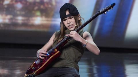 Olly playing a red electric guitar on his knees with a look of deep concentration. He is on stage, and wearing a black back to front baseball cap, a green vest top and black shorts. His hair is light brown and reaches beyond his shoulders.