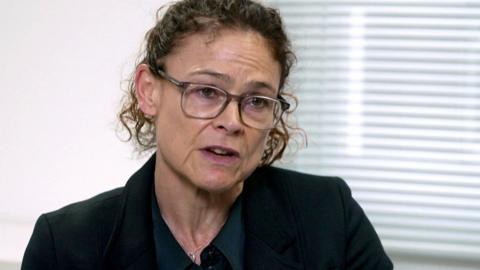 A woman with curly hair and glasses speaks to the camera. She is wearing a balck jacket, and behind her is a white wall and blinds